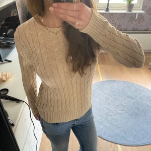 Beige stickad tröja från Polo Ralph Lauren - Säljer en klassisk beige stickad tröja från Polo Ralph Lauren. Tröjan har kabelstickat mönster, rund halsringning och långa ärmar. Perfekt till jeans eller kjol för en stilren look.