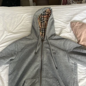 Burberry hoodie - Säljer en grå hoodie från Burberry med dragkedja och huva. Insidan av huvan har det ikoniska Burberry-rutiga fodret. Säljer denna då jag behöver pengar till moppe delar. Skick 10/10 använd relativt fåtal gånger. Kan säljas för 500 vid snabb affär. Mvh