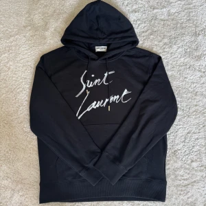 Saint Laurent hoodie - Säljer en svart hoodie från Saint Laurent med vit logga framtill. Tröjan har huva med dragsko och en klassisk känguruficka. Perfekt för en avslappnad och stilren look.
