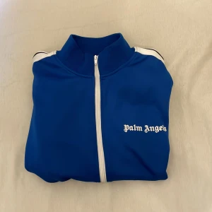 Blå Palm Angels - Säljer en blå track jacket från Palm Angels med vita och svarta ränder längs ärmarna. Jackan har dragkedja framtill och vit logotyp på bröstet. Tillverkad i polyester och har en sportig look.