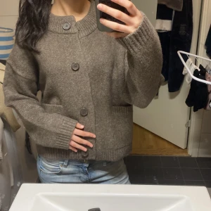 Brun kofta med stora knappar - Säljer en brun, stickad kofta med stora knappar och ribbade muddar. Koftan har en hög krage och är lite oversized i modellen. Perfekt att slänga på sig över en t-shirt eller linne.