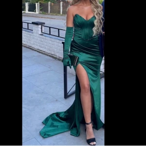 Grön långklänning med slits och handskar - Säljer en elegant grön långklänning med hjärtformad urringning och markerad midja. Klänningen har en hög slits framtill och matchande långa handskar. Perfekt för bal eller fest. Materialet har en glansig finish och klänningen har en figurnära passform.