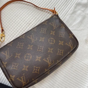 Louis Vuitton monogram axelväska - Säljer min Louis Vuitton pochette med ikoniskt monogrammönster och guldfärgad dragkedja. Väskan har en smal axelrem i läder och är perfekt för att bära det viktigaste. En tidlös accessoar som lyfter varje outfit. Dragkedjan funkar hur bra som helst även den man drar i gått sönder. Finns någon defekt inuti väskan och på väskans handtag annars väldigt fint skick.