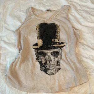 Beige linnetopp med dödskalletryck - Cool beige linnetopp med ett stort tryck av en dödskalle med hög hatt på framsidan. Perfekt för dig som gillar edgy och unika plagg. Tunt och mjukt material, passar bra till jeans eller shorts.