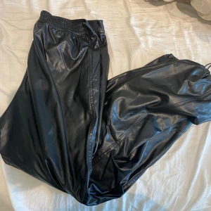 Svarta parachute pants i skinnimitation från H&M - Säljer ett par svarta parachute pants i skinnimitation från H&M med resår i midjan. Byxorna har en ballong passform och är perfekta för att ge en cool look. Materialet är syntetiskt och de är riktigt mjuka och bekväma.