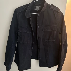 Mörkblå jacka från Crocker - Blå jacka från Crocker i storlek XS. Jackan har klassisk krage, knäppning framtill med stora knappar och två bröstfickor med lock.