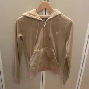 Beige velour hoodie från Juicy Couture - Säljer en beige hoodie i mjuk velour från Juicy Couture. Tröjan har dragkedja, huva med snören och fickor framtill. Ribbad mudd vid ärmslut och nederkant i ljusrosa. Perfekt för en avslappnad och trendig look.