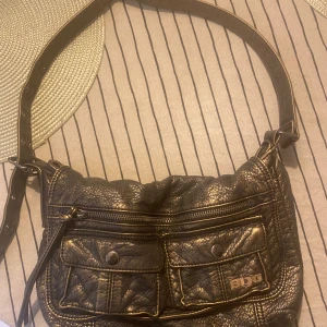 Svart axelväska från BDG - Cool svart axelväska från BDG i skinnimitation med två fickor framtill med knappar och en dragkedja upptill. Väskan har justerbart axelband med nitar och detaljerad struktur. Perfekt för att bära dina viktigaste saker med stil.