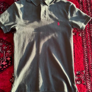 Grön pikétröja från Polo Ralph Lauren - Säljer en klassisk grön pikétröja från Polo Ralph Lauren med korta ärmar och krage. Tröjan har två knappar framtill och en broderad rosa logga på bröstet. Slim fit-modell som passar perfekt till sommaren.