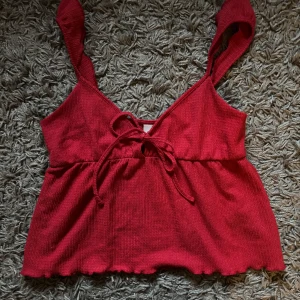Röd croppad topp från Nelly - Säljer en röd croppad topp från Nelly med knyt framtill och breda axelband. Toppen har en lätt rynkad nederkant och är tillverkad i ett ribbat material. Perfekt för varma dagar! Endast använd en gång och säljs på grund av att den är för stor för mig!❤️
