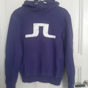Lila hoodie från J.Lindeberg - Säljer en lila hoodie från J.Lindeberg med stor vit logga på bröstet. Tröjan har huva och långa ärmar, perfekt för en avslappnad stil. Mjuk insida och klassisk passform. Snygg och enkel att matcha med jeans eller träningsbyxor.