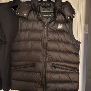 Svart dunväst från BVALDI - Säljer en svart dunväst från BVALDI med avtagbar huva och dragkedja framtill. Västen har två fickor och en inne ficka med dragkedja och en diskret logga på bröstet. Perfekt för lager-på-lager under kyliga dagar. Säljer för den är lite för stor för mig.