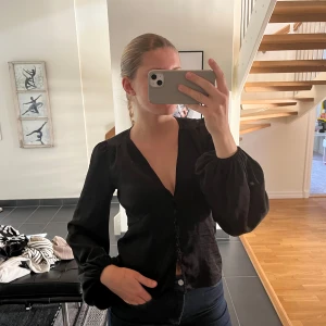 Svart blus med knappar - Säljer en svart blus med v-ringning och knappar framtill. Blusen har långa, puffiga ärmar och är tillverkad i ett glansigt material som ger en elegant känsla. Perfekt att styla med jeans eller kjol.
