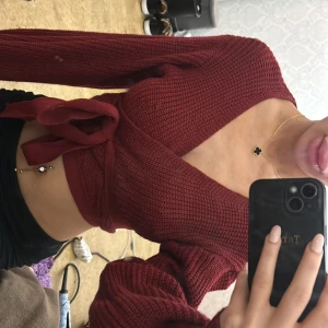 Vinröd omlottopp med knyt - Säljer en vinröd omlottopp med knyt i midjan. Toppen är croppad och har lång ärm. Stickad struktur.