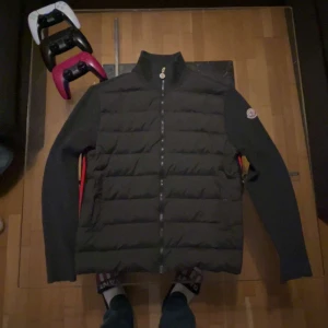 Grå pufferjacka från Moncler - Säljer en svart pufferjacka från Moncler med stickad krage och ärmar. Jackan har dragkedja framtill och Moncler-logga på ärmen. Perfekt för kyliga dagar och har en stilren design.