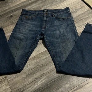 Blå jeans från Hugo Boss - Säljer ett par klassiska blå jeans från Hugo Boss med snygga slitningar och raka ben. Jeansen har fem fickor och detaljer i brunt läder vid bakfickan samt metallmärke fram. Perfekta för en stilren och avslappnad look.