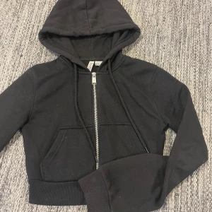 Svart croppad hoodie - Säljer en svart croppad hoodie från H&M. Tröjan har dragkedja framtill, huva med snören och ribbade muddar vid ärmslut och nederkant. Lite fläckar vid ärmarna och jag är osäker på om det går bort i tvätten.