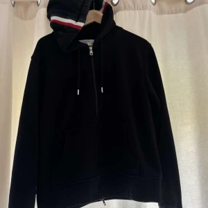 Svart hoodie med dragkedja från Moncler - Svart hoodie från Moncler med hel dragkedja framtill och fickor. Huva med röd, vit och blå randig detalj samt Moncler-logga på ärmen. 