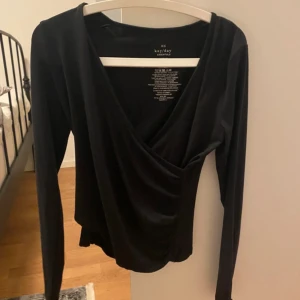 Svart omlottopp från Kay/Day - Säljer en svart omlottopp från Kay/Day i storlek XS. Toppen har lång ärm och djup v-ringning, perfekt för en stilren look. Tillverkad i mjukt material som sitter snyggt på kroppen.