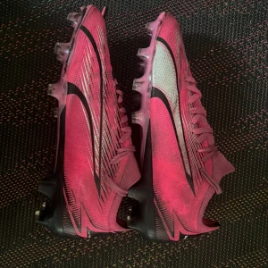 Rosa Nike fotbollsskor med svarta detaljer - Säljer ett par rosa Nike fotbollsskor med svarta och vita detaljer. Skorna har snörning och mönstrad ovansida samt dobbar för bra grepp på planen. Perfekta för dig som vill sticka ut på fotbollsplanen.