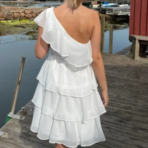 Vit volangklänning från Vila - Superfin vit klänning från Vila med volanger och omlottkänsla. Klänningen har en axelbandslös design och knytdetalj i midjan. Perfekt för studenten eller skolavslutning! Fråga vid funderingar🩷