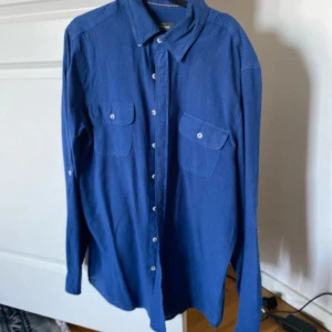 Blå linneskjorta från Dressmann - Säljer en stilren blå skjorta från Dressmann i linnemix. Skjortan har långa ärmar, två bröstfickor med knappar och klassisk krage. Perfekt för dig som gillar enkel och snygg stil.