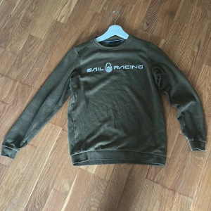 Olivgrön sweatshirt från Sail Racing - Säljer en olivgrön sweatshirt från Sail Racing med vit logga tryckt på bröstet. Tröjan har rund halsringning och långa ärmar. Perfekt för en avslappnad stil och enkel att matcha med jeans eller joggers.