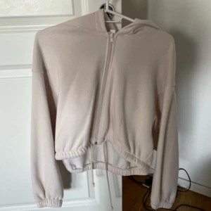 Ljusbeige hoodie med dragkedja - Säljer en ljusbeige hoodie med dragkedja och huva. Jackan har långa ärmar, resår i midjan och vid ärmsluten samt dragsko nertill. Perfekt för en avslappnad stil och enkel att matcha med olika outfits.