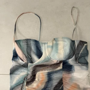 Flerfärgad omlottklänning från Gina Tricot - Supersnygg kort topp från Gina Tricot med abstrakt mönster i blått, vitt, beige och orange. Toppen har smala axelband, omlott-effekt och lätt rynkade detaljer framtill. Perfekt för dig som vill sticka ut lite extra!