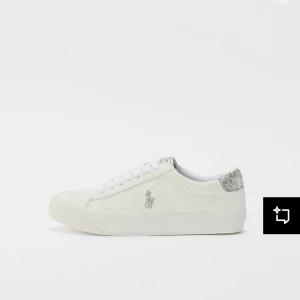 Vita sneakers från Polo Ralph Lauren med glittrig detalj - Snygga vita sneakers från Polo Ralph Lauren med glittrig silverdetalj på hälen och klassisk logga på sidan. Skorna har snörning och en låg, platt sula. Perfekta för en stilren och trendig look.