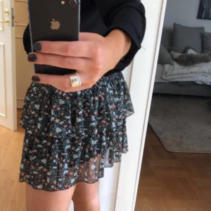 Blommig volangkjol i svart och grönt - Säljer en kort blommig kjol med volanger. Kjolen har svart bas med små blommor i grönt, vitt och rosa. Den har en luftig och lätt känsla, perfekt för varmare dagar. Resår i midjan för skön passform.