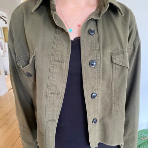 Grön jacka  - Säljer en olivgrön overshirt med stora svarta knappar och två bröstfickor. Jackan har en rak, lite boxig passform och är tillverkad i ett kraftigare bomullstyg. Perfekt att slänga över en t-shirt eller linne.