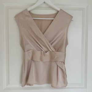 Beige omlottopp - Säljer en stilren beige omlottopp med bred midjedetalj och ärmlös design. Toppen har en v-ringad omlottfront och är tillverkad i ett mjukt, glansigt material som ger en elegant känsla.