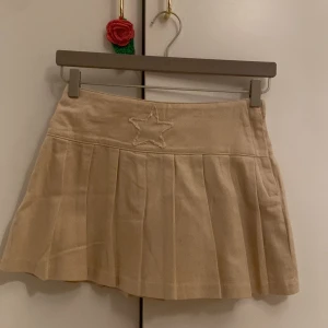 Beige kortkjol med stjärndetalj - Säljer en beige kortkjol med veck och en broderad stjärna framtill i midjan. Kjolen har en klassisk, lätt utställd form och är perfekt för dig som gillar en enkel men söt stil.