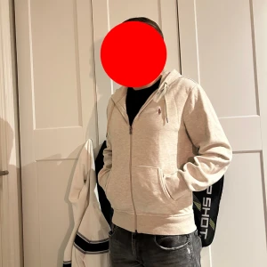Ljusgrå hoodie med dragkedja från Ralph Lauren - Säljer en ljusgrå hoodie från Ralph Lauren med dragkedja framtill och huva. Tröjan har två fickor på magen och en liten broderad logga på bröstet. Perfekt för en avslappnad stil.