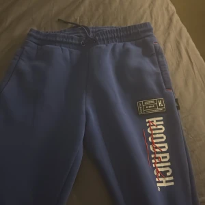 Blå mjukisbyxor från Hoodrich - Säljer ett par blå mjukisbyxor från Hoodrich med tryckta loggor och detaljer i vitt och rött. Byxorna har resår och snörning i midjan samt muddar vid bensluten. Perfekta för en avslappnad stil. 