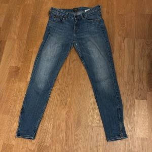 Blå jeansbyxor från Lee med dragkedja - Säljer ett par klassiska blå jeans från Lee med fem fickor och dragkedja. Byxorna har en rak passform och subtila slitningar. Dem har stretch är lågmidjade.