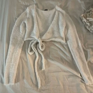Vit croppad kofta från NA-KD - Supermjuk och fluffig vit kofta från NA-KD. Modellen är croppad med långa ärmar och knyt framtill. Perfekt att slänga över en topp för en mysig och trendig look.
