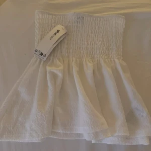 Vit bandeau tubtopp från Gina Tricot - Supersöt vit bandeau tubtopp från Gina Tricot med smockad överdel och volangig nederkant. Toppen är ärmlös och har en luftig, lätt struktur i tyget som ger en fräsch känsla. Perfekt för varma dagar och enkel att styla med jeans eller kjol.