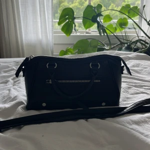 Svart handväska från Primark - Snygg svart handväska från Primark med stilrena silverdetaljer och dragkedja framtill. Väskan har både handtag och lång axelrem, perfekt för att bära på olika sätt. Tillverkad i syntetmaterial med ett rymligt fack och innerficka med dragkedja.