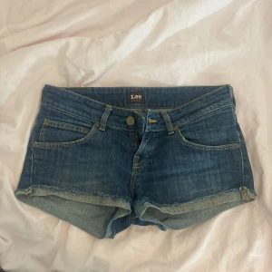 Blå jeansshorts från Lee - Snygga blå jeansshorts från Lee med råa kanter och klassiska fem fickor. Shortsen har normal passform och lowwaist, perfekt för sommardagar. Tidlös denimlook med Lee-logga bak i midjan och slitna detaljer för en avslappnad vibe.