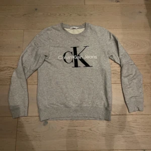 Grå Calvin Klein Jeans sweatshirt - Snygg grå sweatshirt från Calvin Klein Jeans med stort svart CK-tryck på bröstet. Tröjan har rund hals, långa ärmar och ribbade muddar. Perfekt för en chill och stilren look.