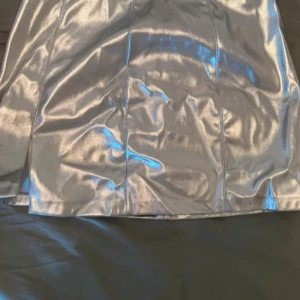 Silvrig kortkjol från Pimkie - Supersnygg silvrig kortkjol från Pimkie i glansigt material. Kjolen har en rak passform och dragkedja bak för enkel påklädning. Perfekt för dig som vill sticka ut med en futuristisk vibe och metallic-look.