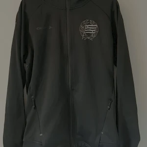 Svart zip från Craft, storlek S - Svart vindjacka från Craft i återvunnen polyester. Jackan har hel dragkedja, huva och två fickor med dragkedja. På bröstet finns en vit logga med lagerkrans och flagga. Perfekt för dig som gillar sportig och clean stil.