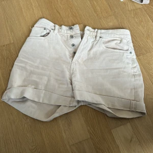 Beige jeansshorts från lager 157, storlek M - Snygga beige jeansshorts från lager 157 i storlek M. Modellen är högmidjad med klassiska fem fickor, uppvikta benslut och knäppning med flera knappar framtill. Perfekta för varma dagar och enkel att matcha med allt.