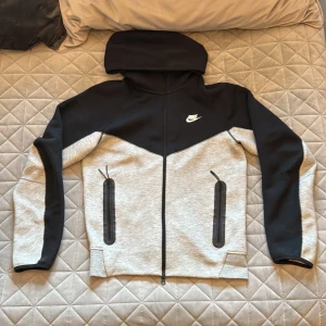 Nike Tech Fleece hoodie svart/grå - ⚠️Diskuterbart pris ⚠️Nike Tech Fleece hoodie i svart och ljusgrått med hel dragkedja och två stora fickor med svarta dragkedjor. Snygg sportig look med huva och klassisk Nike-logga på bröstet. Perfekt för varje dags användning eller träning. Skicka om det finns minsta lilla fundering.