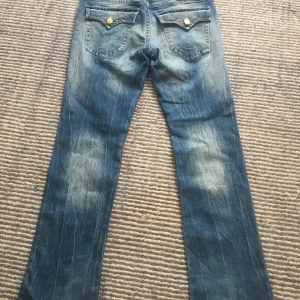 True Religion Ricky Relaxed Straight Jeans - Säljer ett par fire blå True Religion Ricky Relaxed Straight jeans med slitningar och hål på framsidan. Klassiska bakfickor med lock och guldiga knappar. Jeansen har coolt tvättad look, perfekta för dig som gillar streetstyle.