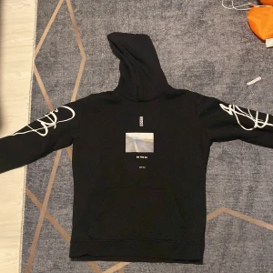 Svart hoodie från Hugo Boss  - Svart hoodie från Hugo Boss i storlek L/XL med coolt tryck av en väg och texten 'ON THE GO' på bröstet. Vita grafiska detaljer på båda ärmarna och klassisk huva. Perfekt för dig som gillar streetstyle och vill sticka ut.