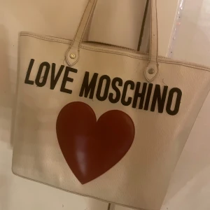 Love Moschino shoppingväska med hjärta - Stor och rymlig shoppingväska från Love Moschino i beige skinnimitation med svart text och ett stort rött hjärta på framsidan. Väskan har två långa handtag och dragkedja upptill. Perfekt för dig som vill sticka ut med en ikonisk design. Väskan är nästan oanvänd och är väldigt rymlig💕originalpris: 3599