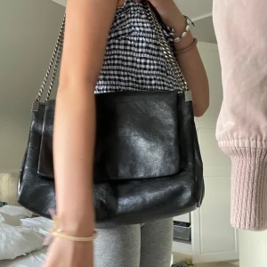 Svart axelväska med kedjerem - Stilren svart axelväska i skinnimitation köpt på Zara. Väskan har en enkel, fyrkantig form och passar perfekt till en trendig vardagslook. Rymmer allt du behöver och ger en edgy touch till outfiten. har börjat rosta pyttelite på ena kedjan, skriv för bilder.